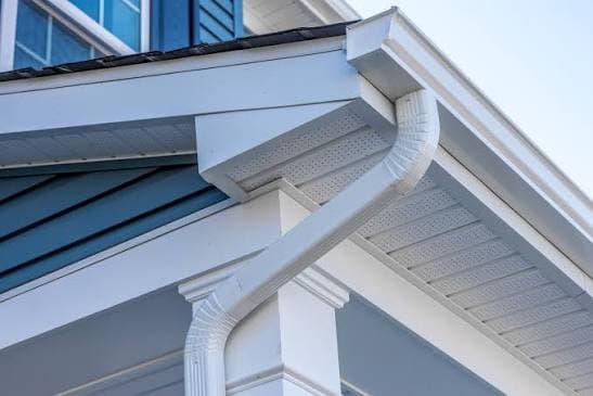 Close-up of a new white seamless gutter and downspout at a roof corner, above vented white soffit